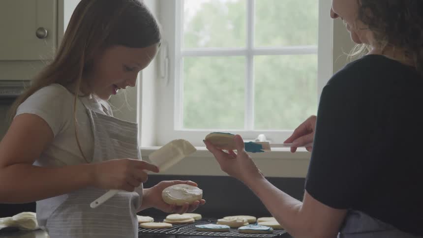 A mother and daughter smile as they frost sugar cookies in their kitchen