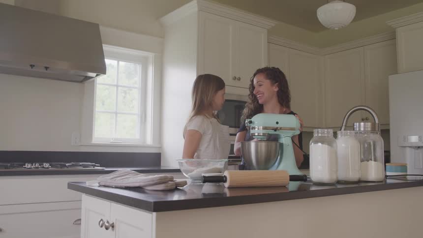 A woman and daughter stand in the kitchen with a mixer and baking ingredients