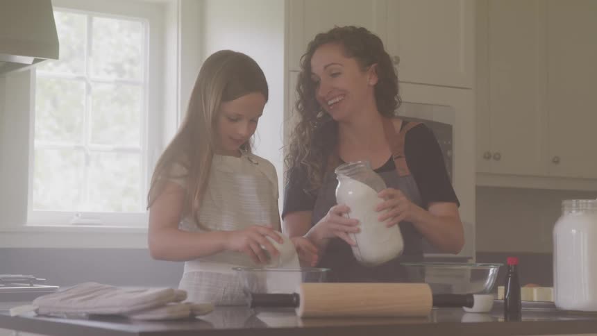 A woman pours sugar from a jar into a small bowl being held by her daughter as they cook together in a kitchen