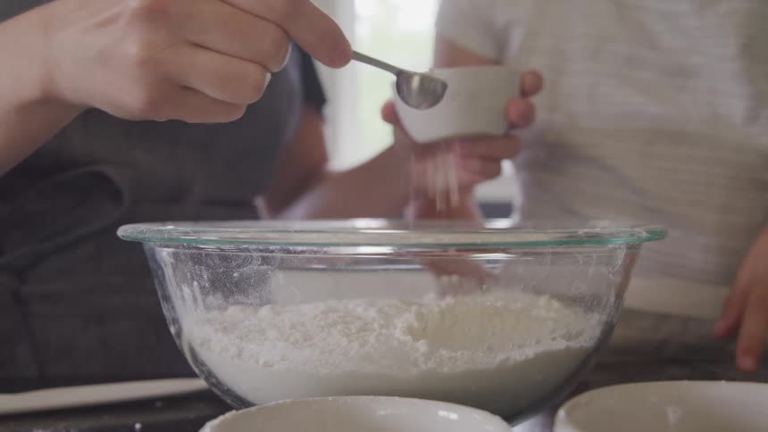 Woman cooking at home uses a spoon to pour salt on a glass mixing bowl filled with white flour