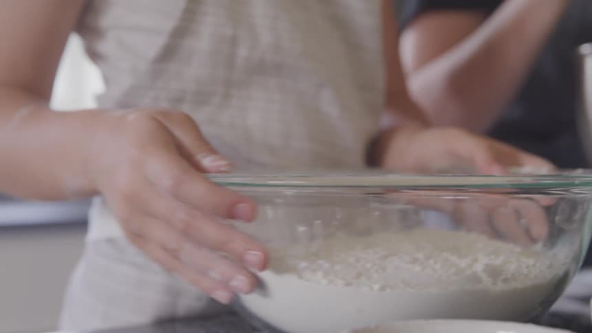 A young girl holds the sides of a glass mixing bowl filled with flour as she helps her mother cook at home