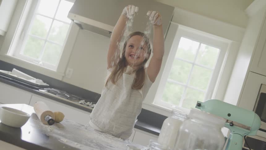 A young girl stands at the kitchen counter and drops flour on it as she bakes
