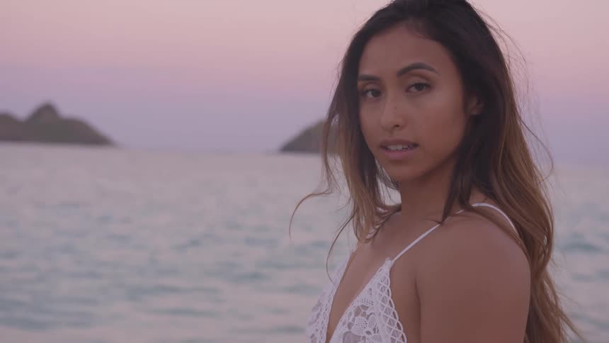 A young woman looks into the camera while standing on the coast at sunset