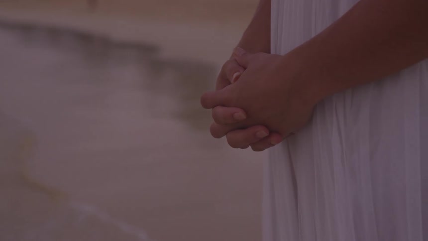 A woman in a white gown clasps her hands in front of her as she stands on a beach