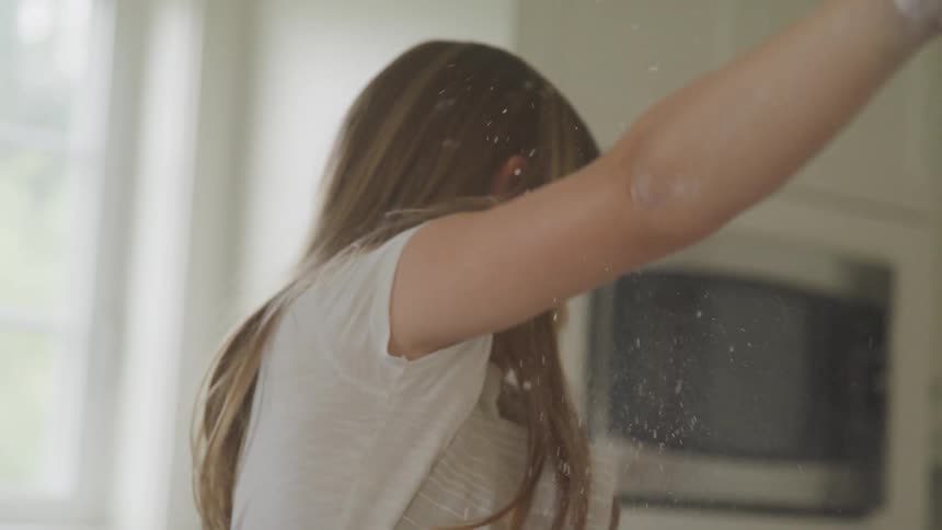 A girl in a kitchen smiling and throwing flour with a window in the background