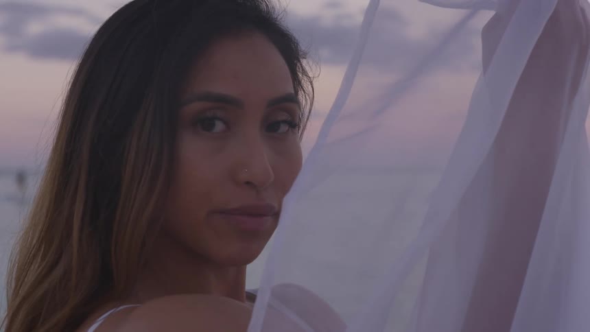 Woman holding white veil in hand stands in front of ocean at dusk