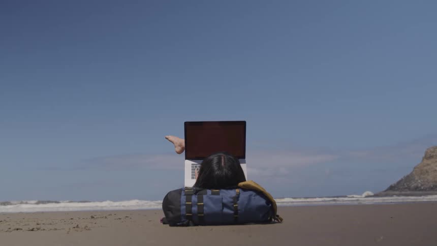 A woman lies with her head resting on her backpack as she uses a laptop on a beach on a sunny day