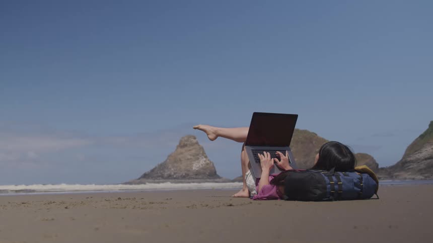 A woman lies down and uses her laptop on the beach