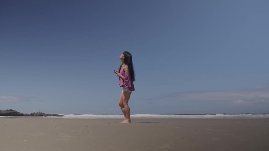 A teenage girl holding her cell phone dances on the beach