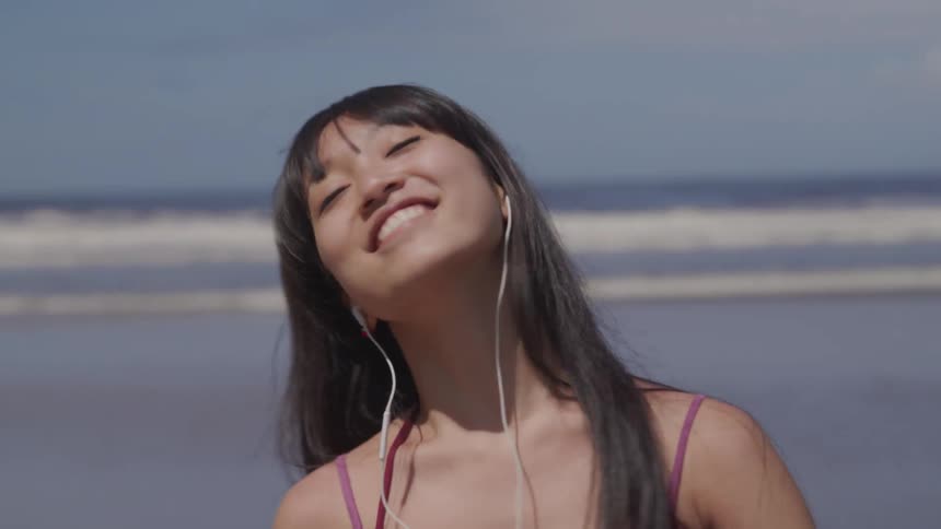 Woman smiles and twirls as she walks across beach listening to music on mobile phone via earbuds