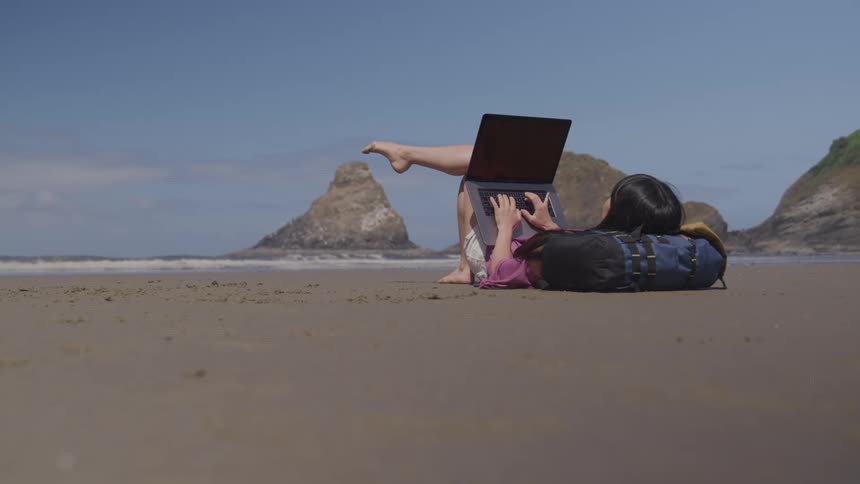 A woman lays on the beach with her laptop propped up on her legs working