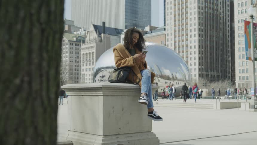 A woman sits on a small city pillar looking at her cell phone