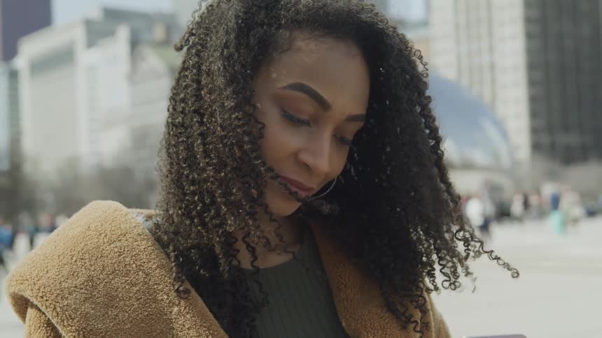 Black woman with curly hair hoop earrings and a brown coat looks at her smartphone
