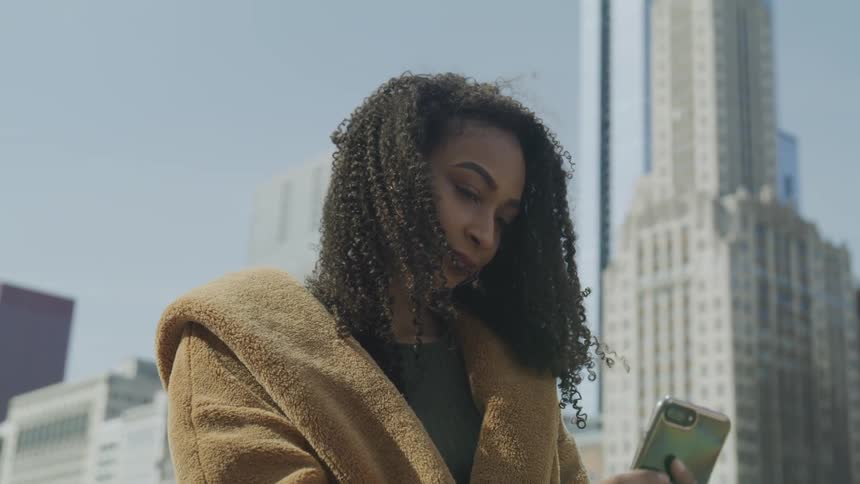 A woman looks at her cell phone with large city buildings behind her