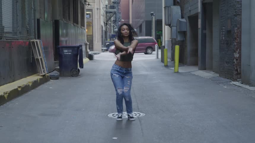 A young woman wearing headphones is dancing outdoors in an alleyway in a city