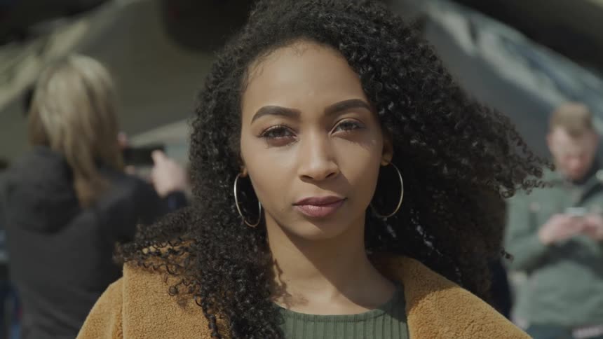 Black woman with curly hair hoop earrings and a brown coat stares at the camera