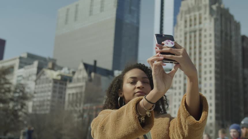 A woman holds up her cell phone to take a selfie as shes standing with large city buildings in the background