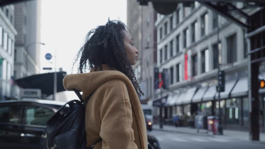 A woman walks across a city street wearing a coat and back pack style purse