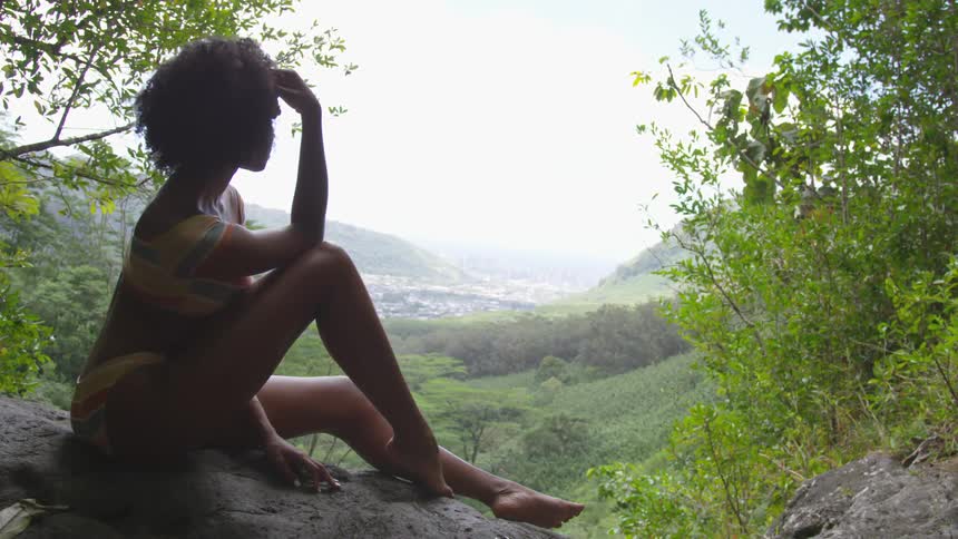 Woman sits on ground wearing bikini looking through valley past town toward horizon
