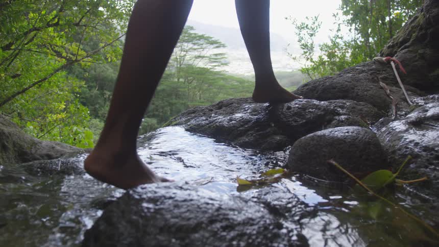 The bare legs and feet of a person walking on rocks across a flowing hilltop stream