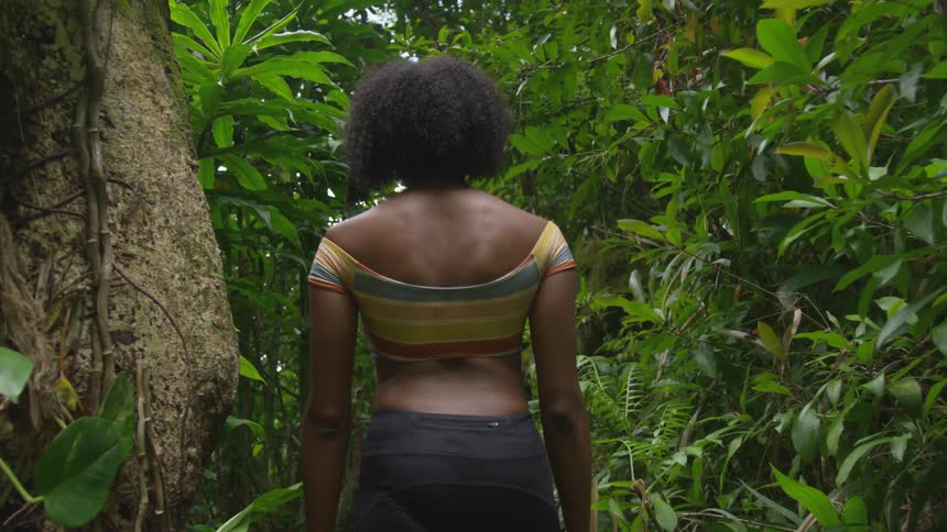 A black woman walks through the thick vegetation of a forest during the day