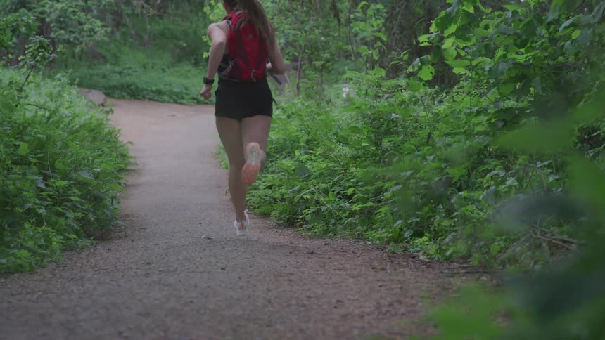 Female wearing backpack runs along a dirt trail in a wooded area for exercise