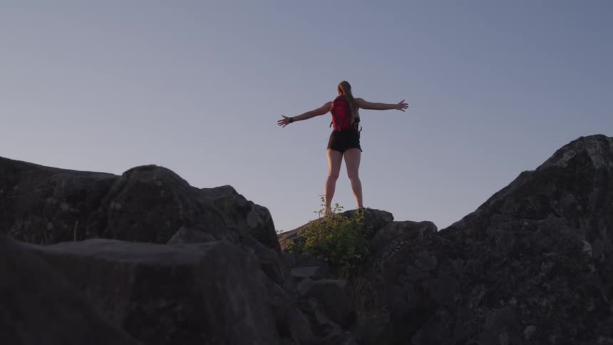A woman standing on a cliff stretches her arms up in the air