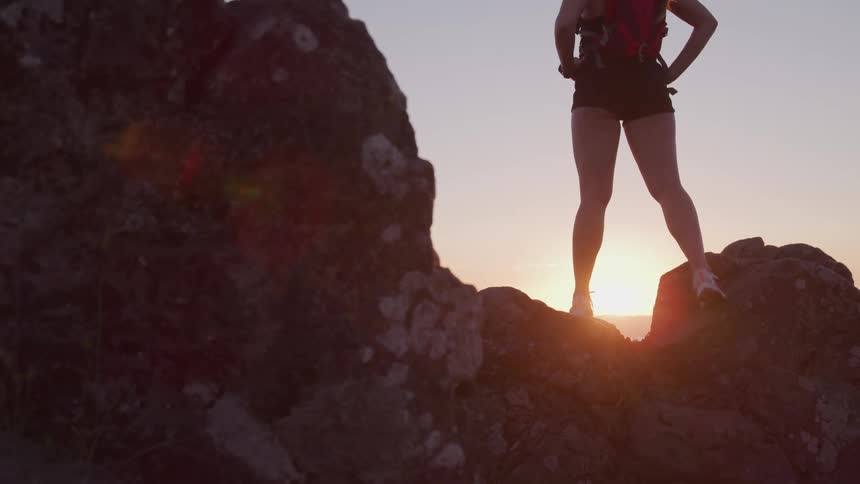 Woman with shorts a red backpack and a pair of shoes stands on rocks and watches the sunset