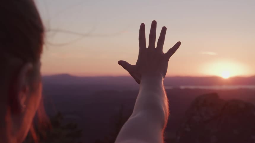 A woman standing outdoors at high altitude reaches her hand out towards the rising sun on the horizon and slowly curls her finger into a fist