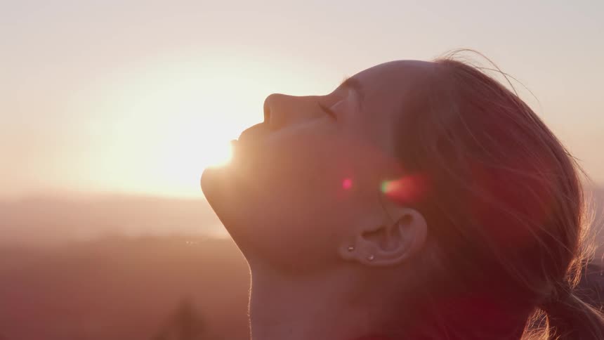 A young woman standing outdoors at sunset closes her eyes as she raises her face towards the sky
