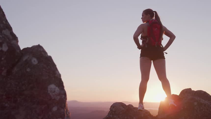A female hiker stands atop a a rock formation on a mountain with her hands on her hips as she looks out at the landscape below