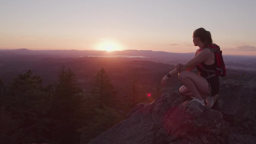 A young woman kneels on the cliff of a mountain and looks out at the sun peeking over the horizon