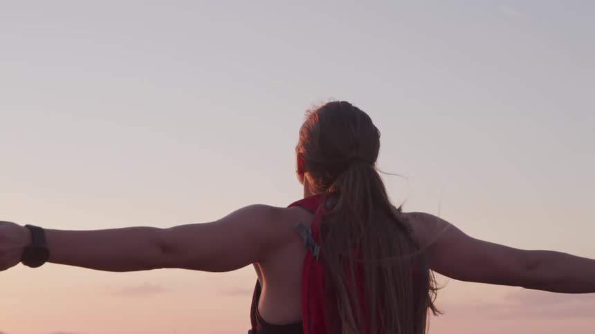 A woman raises her arms out to her sides as she looks at the setting sun