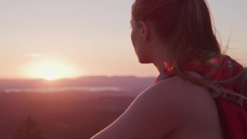 Woman wearing backpack looks toward horizon at sunset