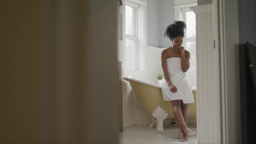 A black woman wearing a towel sits on the edge of a bathtub while lost in thought