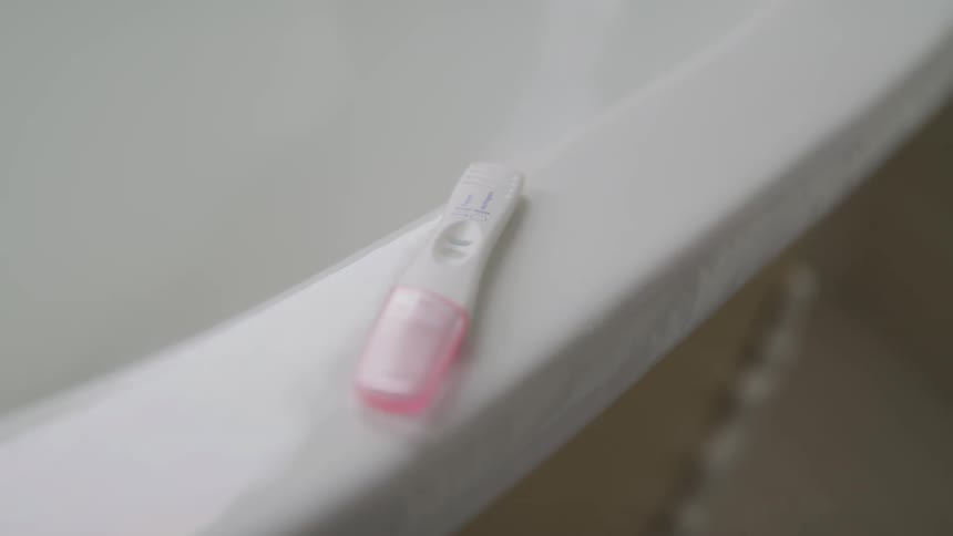 A pregnancy test with a pink cap sits on the lip of a white bathtub