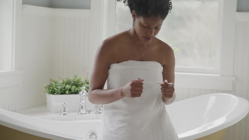 A young black woman sitting on the edge of a bath tub with a towel wrapped around her body somberly checks a pregnancy test