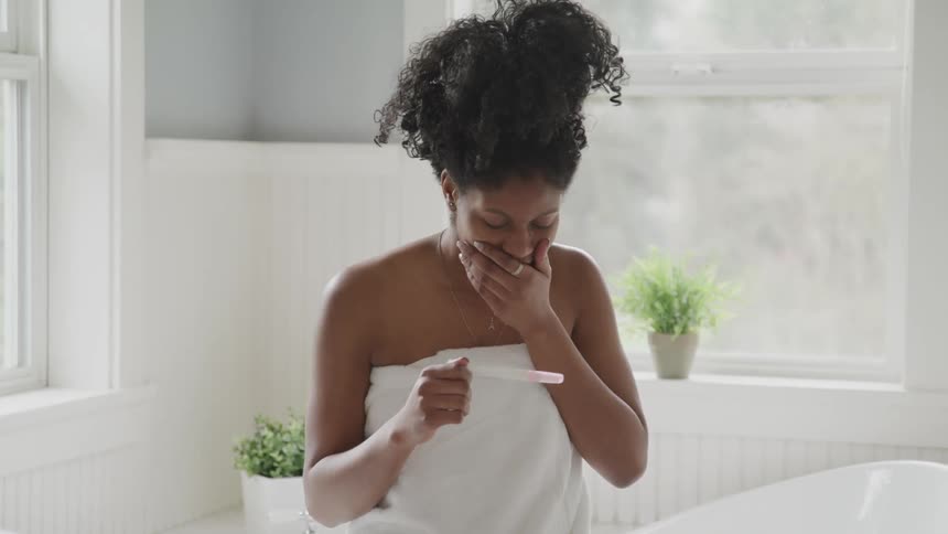 A young woman sitting in her bathroom looks at a pregnancy test and covers her mouth as she smiles