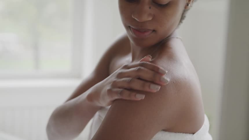A woman sitting in a bathroom with a towel wrapped around her body rubs her shoulder with her hand