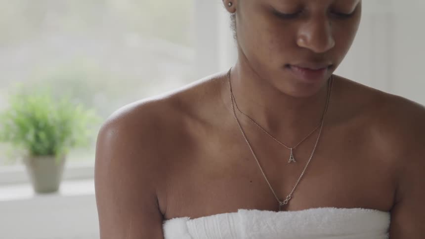 A young black woman wearing a towel rubs cream on her arms