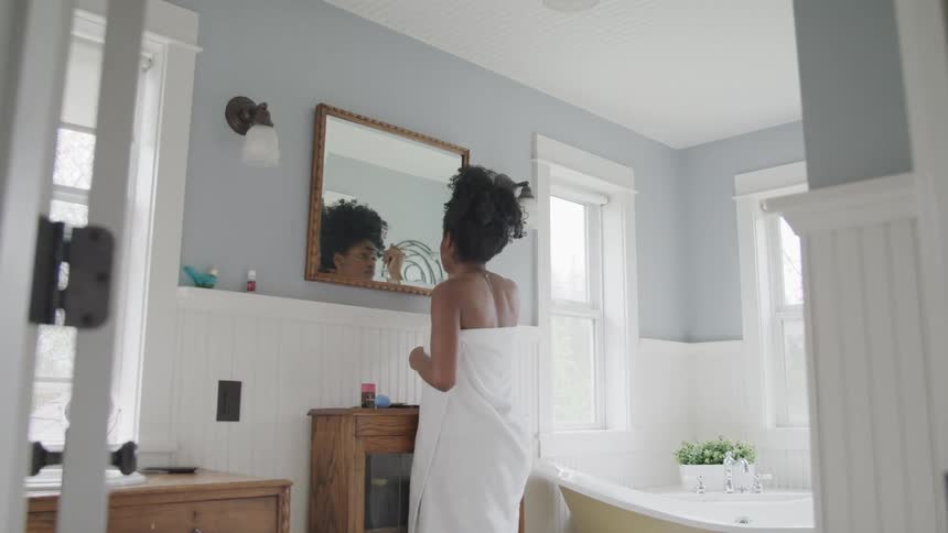 A woman standing in front of a mirror in her bathroom puts make up