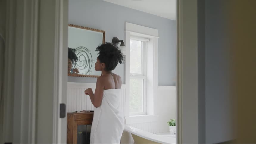 A young black woman standing in front of a mirror at home puts makeup on her face