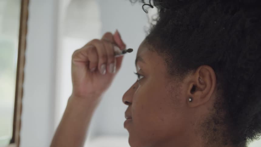 A black woman applies makeup on her eyebrow in front of a mirror