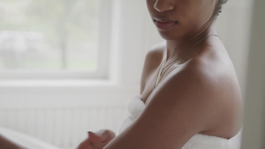 A woman with necklaces wrapped in a towel rubbing lotion on her skin