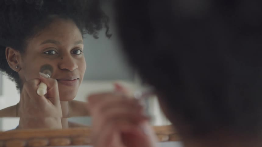 A young black woman applies makeup to her face while looking into a mirror