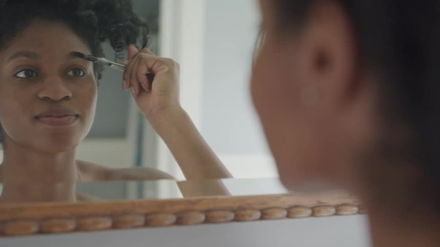 Young woman applies makeup on her eyebrows with a brush in front of the bathroom mirror