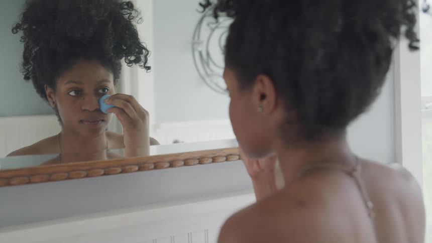 A beautiful woman with natural hair piled on her head pats her face with an egg shaped makeup sponge
