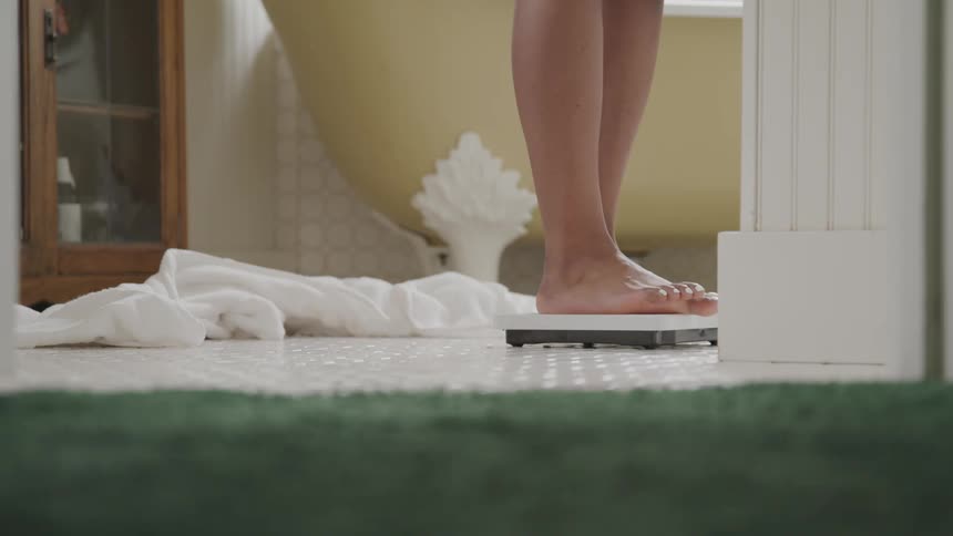 A woman removes a towel from around her body and steps onto a scale to check her body weight
