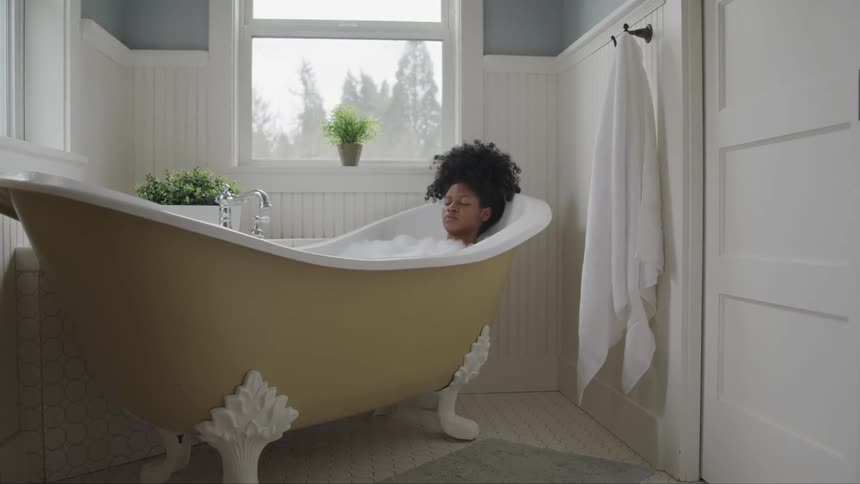 A young black woman sits back with her eyes closed in her bath tub as sunlight shines in through the window