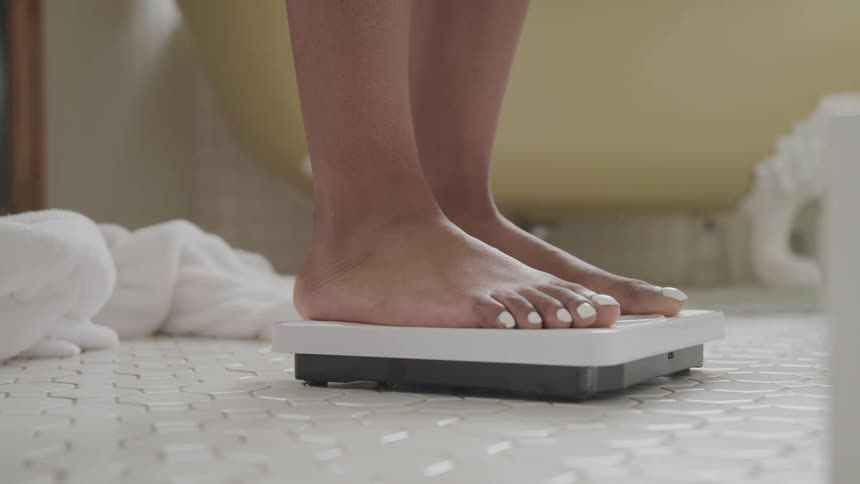 A woman takes off her white bath robe and steps on a scale in her bathroom